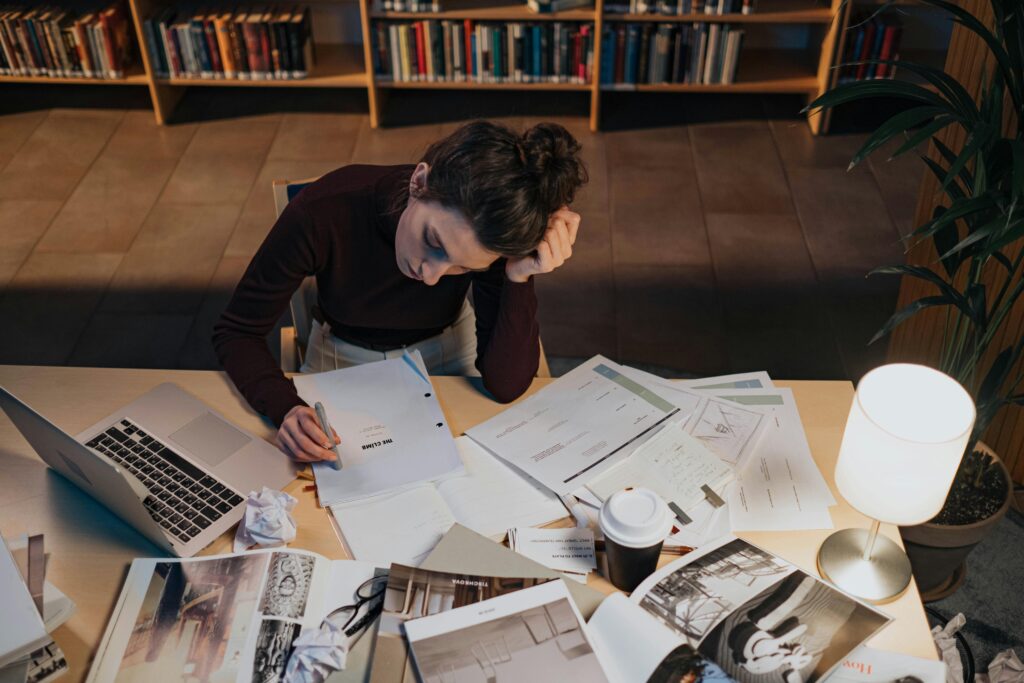 Woman studying at desk with documents, laptop, and coffee in a cozy library setting.